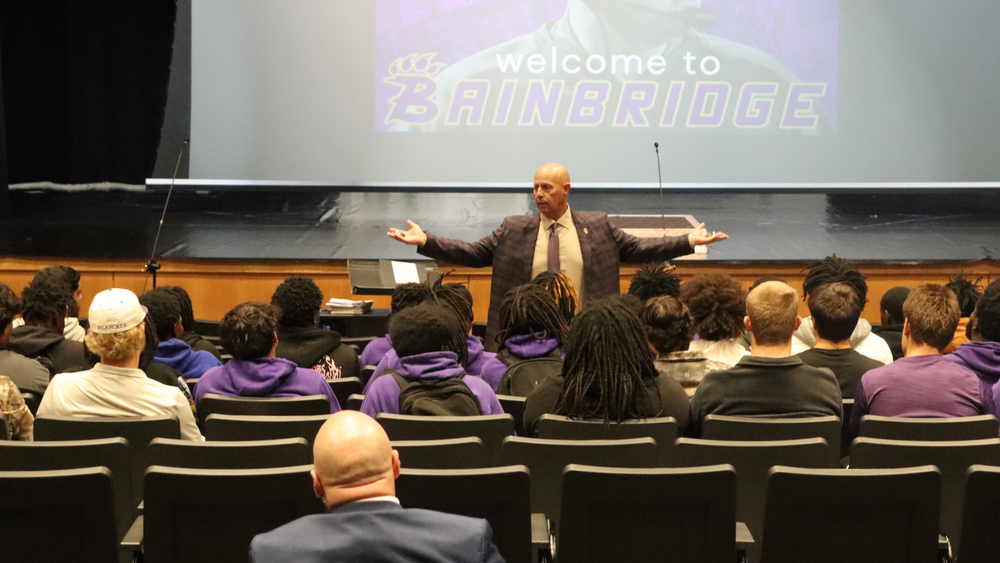 A speaker addresses a seated group of students in an auditorium with a ‘Welcome to Bainbridge’ screen behind him