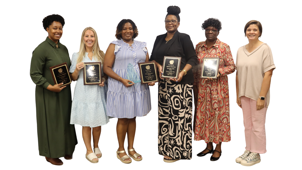 Six women standing side by side, five holding plaques for Teacher of the Year awards. They are smiling and dressed in professional and semi-formal attire, standing against a white background