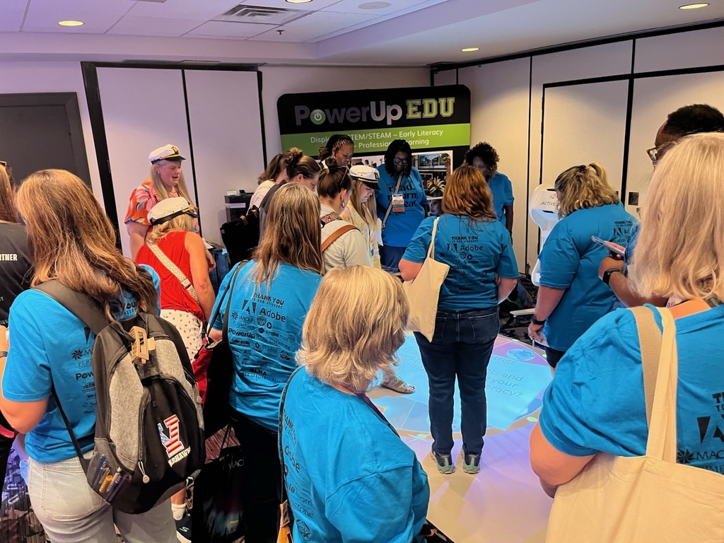 : A group of people in blue shirts gather around an interactive floor display at a literacy and STEAM education booth.