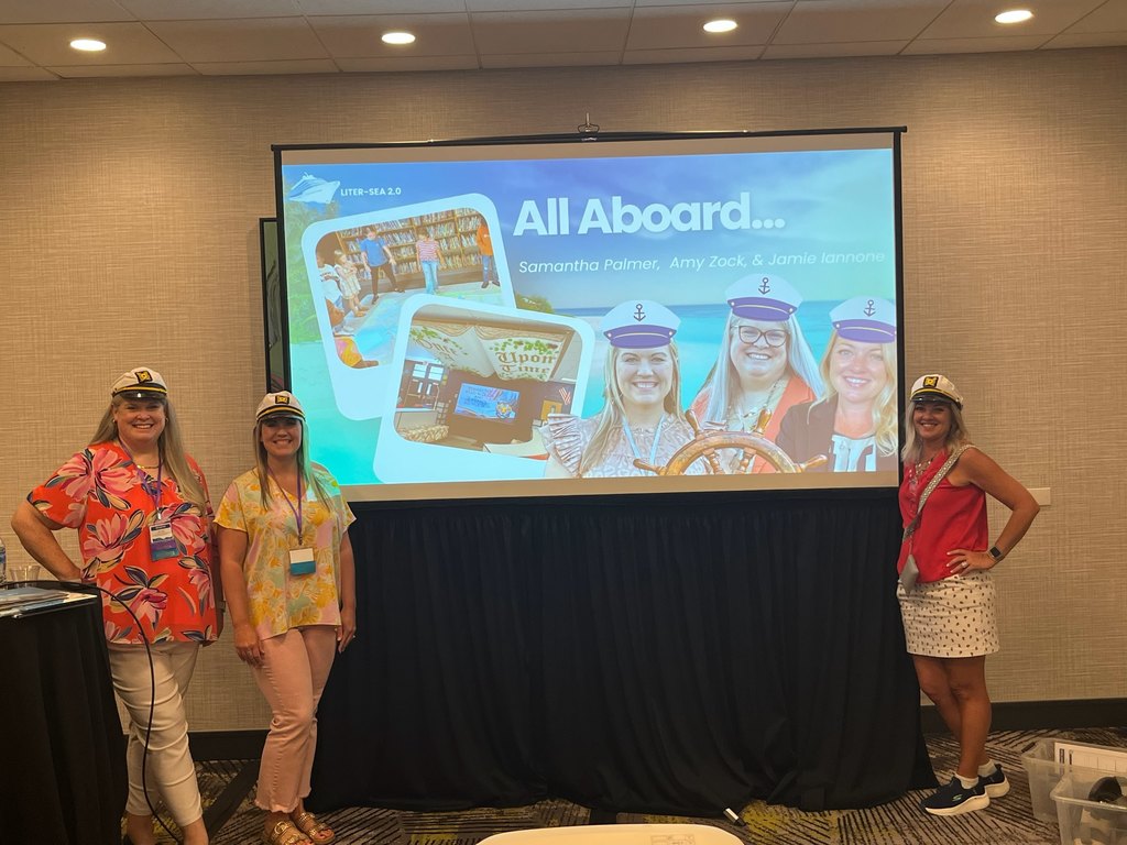 women wearing sailor hats pose in front of a presentation screen in a conference room. The screen displays a colorful slide titled “All Aboard…” featuring photos of a library-themed setting and three women in sailor hats. The presenters appear to be introducing or discussing a project called "LITER-SEA 2.0." The women are smiling and dressed in casual, summery conference attire.