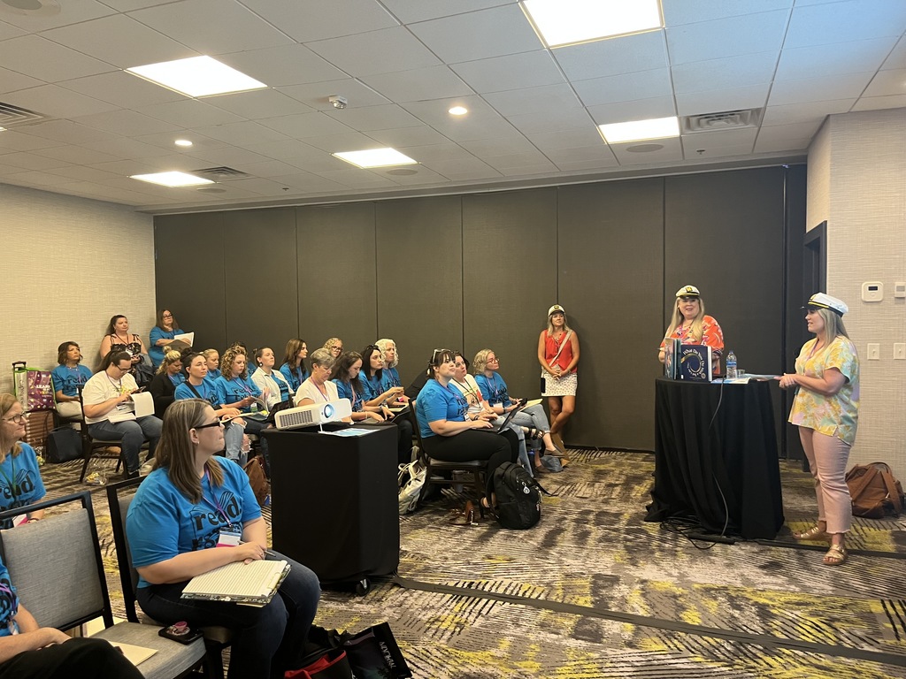 Presenters in sailor hats speak to a seated audience wearing blue shirts during an educational conference session.