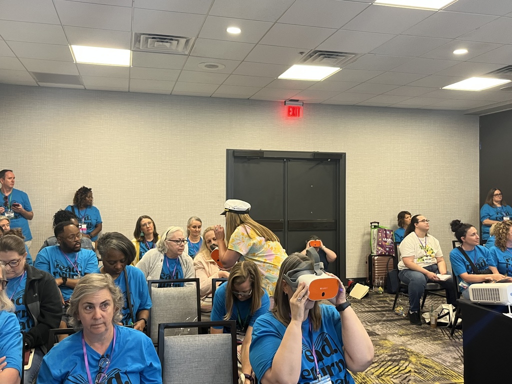 Conference attendees wearing blue shirts explore virtual reality headsets while seated in a session room.
