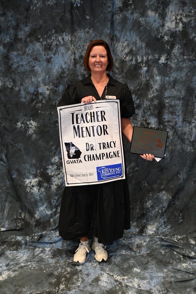Dr. Tracy Champagne stands smiling in front of a mottled backdrop, holding a plaque and a sign that reads "2025 Teacher Mentor – Dr. Tracy Champagne – GVATA," recognizing her as GVATA Teacher Mentor of the Year.