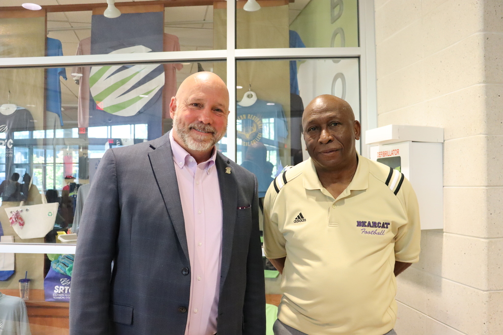 Two men pose for a photo inside the event; one wears a suit and the other a Bearcat Football shirt.