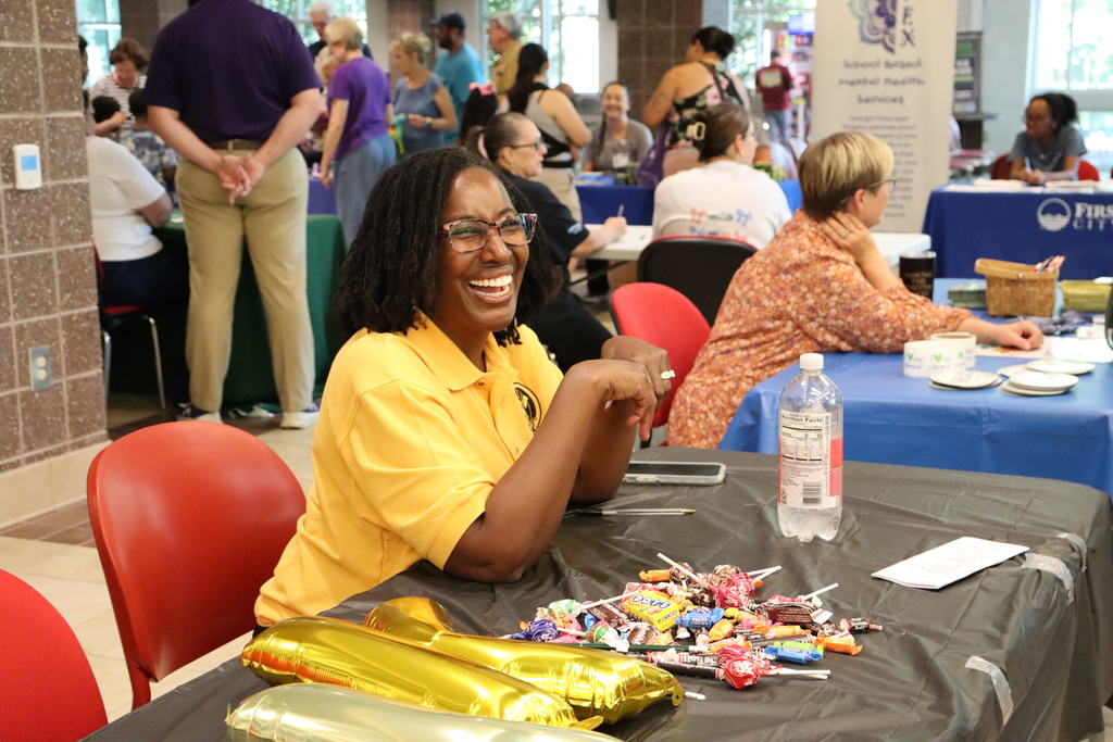 A smiling woman in a yellow shirt sits at a table with candy and gold balloon letters.