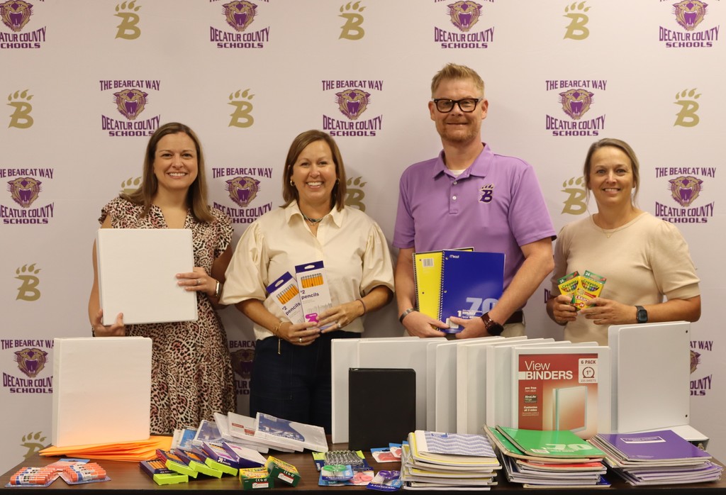 Four adults stand smiling in front of a Decatur County Schools backdrop, holding donated school supplies such as binders, notebooks, pencils, and crayons, with a table full of additional supplies in front of them