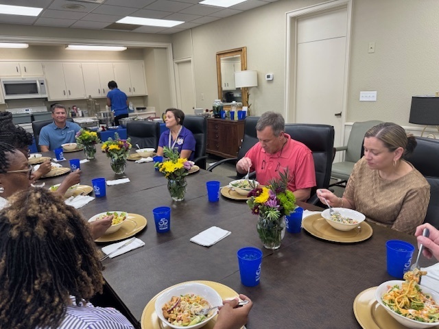 Teachers and staff are seated around a conference table enjoying lunch, with floral centerpieces and blue cups on the table.