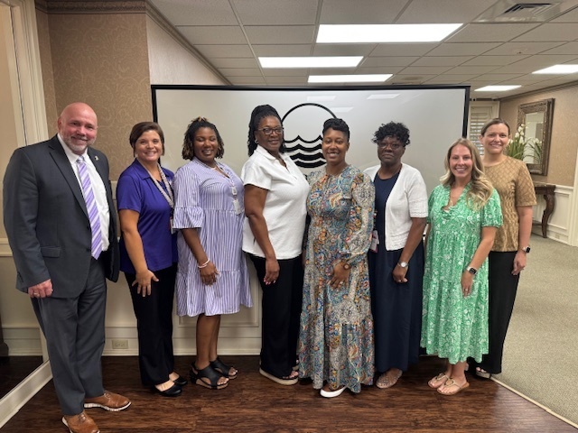 Group photo of the Teachers of the Year standing with Dr. Boyd English in front of a First Port City Bank logo. Everyone is smiling and dressed professionally.