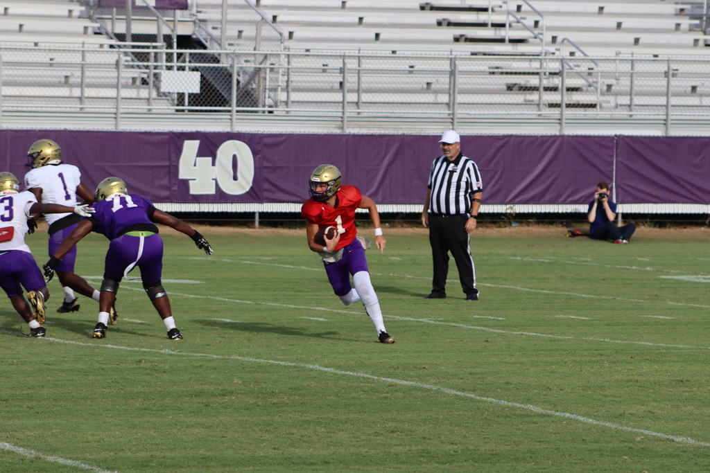 A Bainbridge High School quarterback wearing a red jersey and gold helmet runs with the football during a scrimmage as teammates block and a referee watches nearby.
