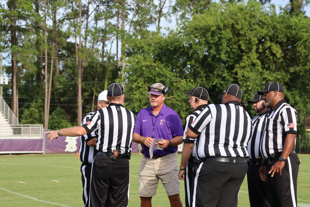 A Bainbridge coach in a purple shirt and visor meets with a group of referees on the football field before the game, surrounded by trees and stadium seating in the background.