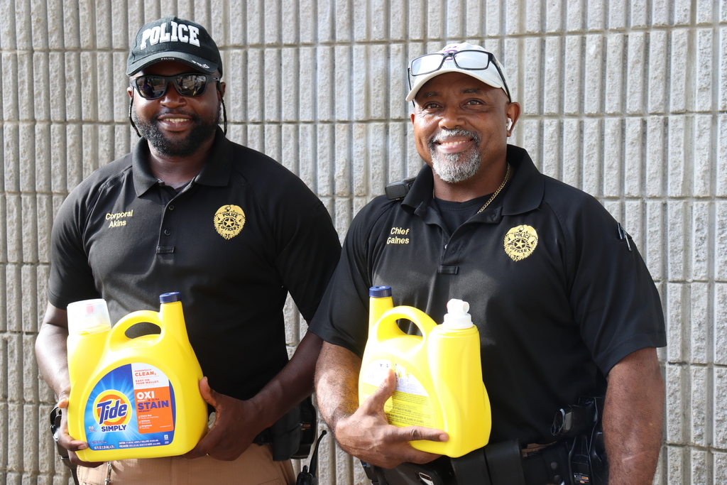Two Bainbridge Public Safety officers, Corporal Akins and Chief Gaines, smile while holding donated bottles of OxiClean and Tide detergent, standing in front of a textured wall.