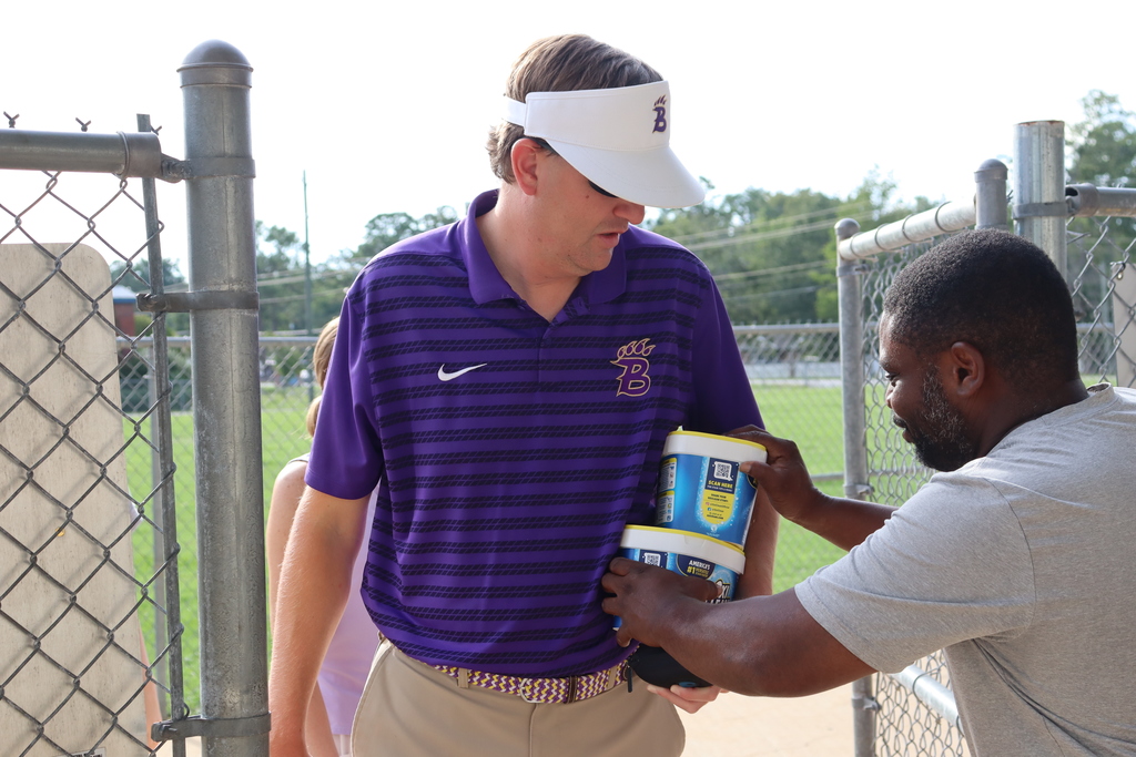 A man hands multiple containers of OxiClean to a Bainbridge staff member wearing a purple Bearcat polo and white visor at a stadium entrance.