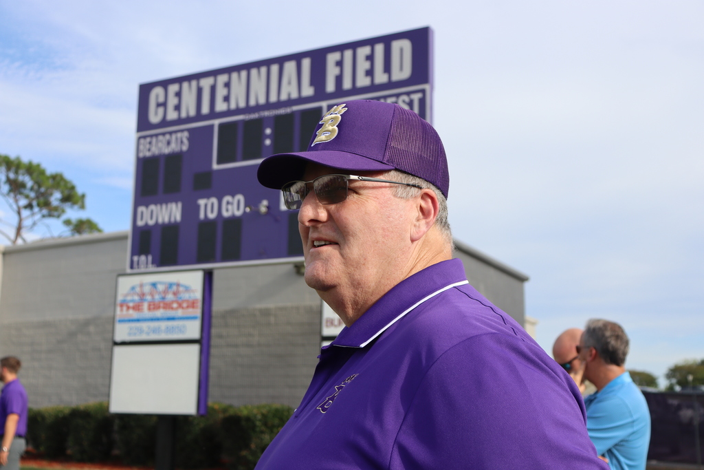  A man in a purple Bainbridge Bearcats polo and hat stands in front of the scoreboard at Centennial Field, smiling as he looks toward the field.