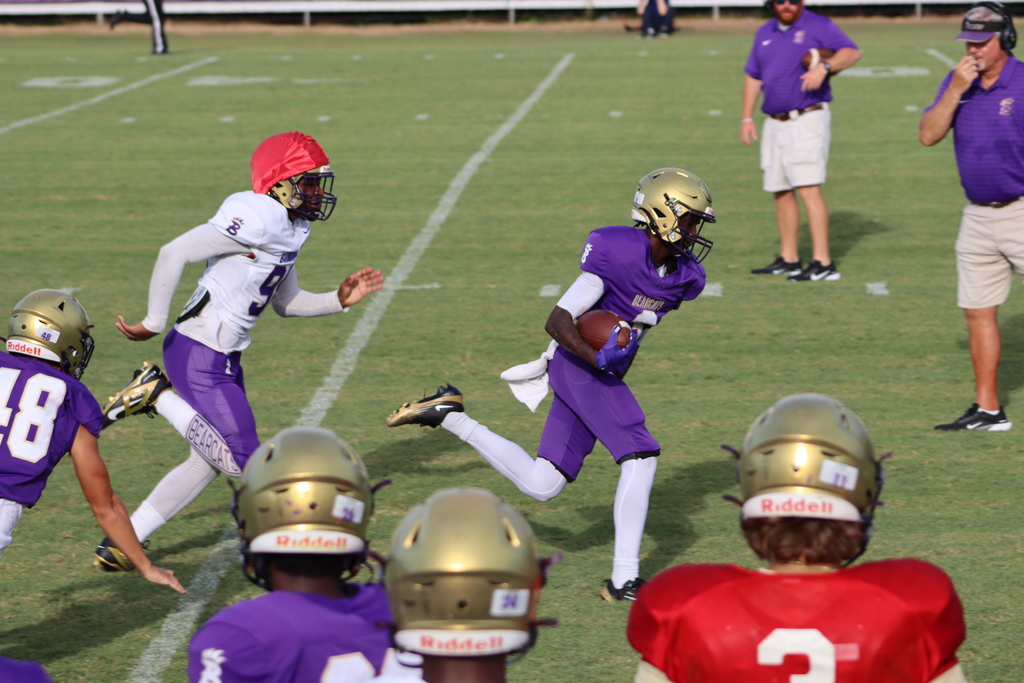  Bainbridge High School football players scrimmaging on the field, with a player in a purple uniform running the ball as teammates and coaches look on.