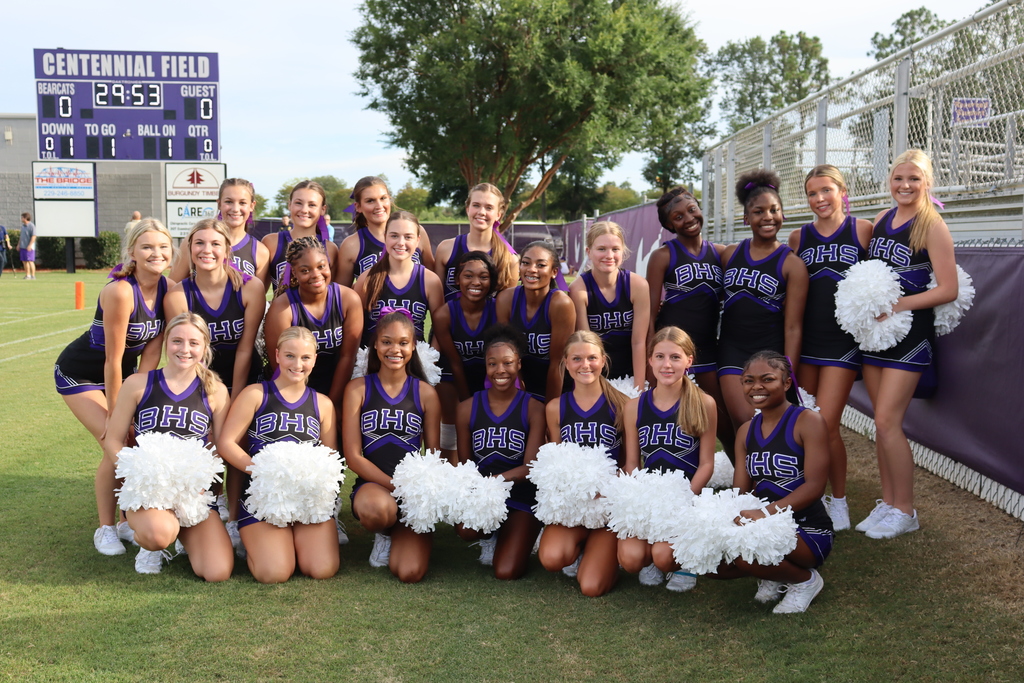 The Bainbridge High School cheerleading squad poses together in uniform on the football field in front of the Centennial Field scoreboard, holding white pom-poms and smiling for the camera.