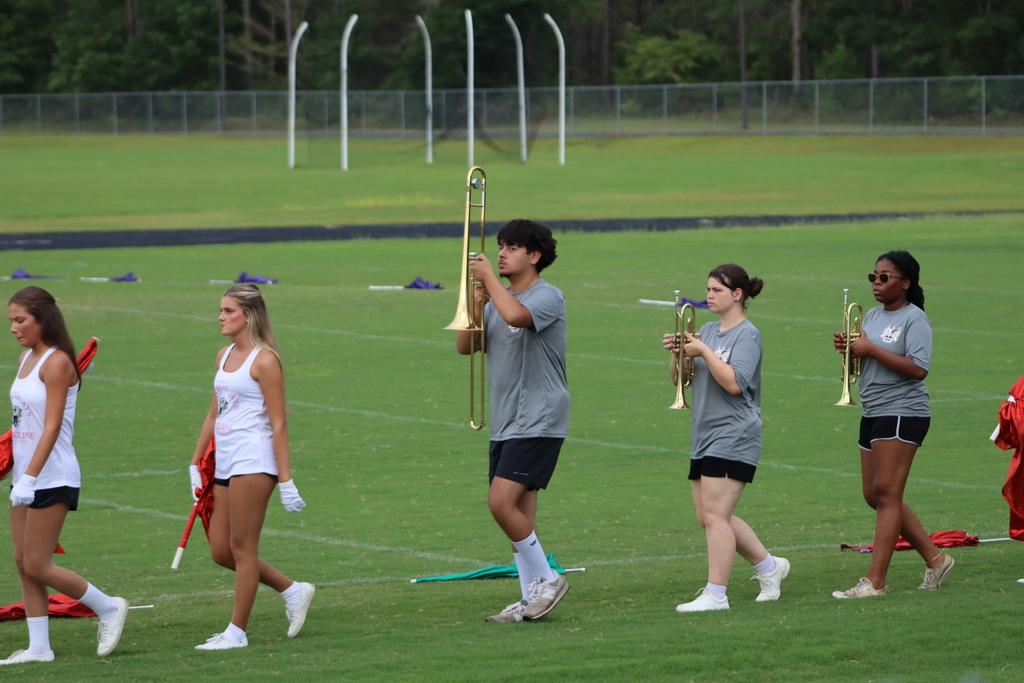 Band and danceline members march in formation across the field; two trumpet players and a trombone player are surrounded by danceline members holding red flags