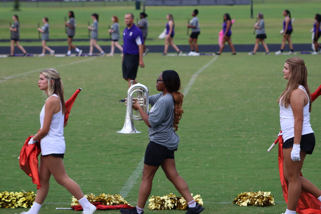 Three students walk across the football field — two members of the danceline holding red flags and one band member carrying a silver marching horn, with other band and auxiliary members in the background.