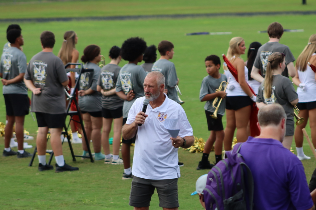 A band director holds a microphone and speaks during Family & Friends Night while students in gray shirts and danceline uniforms gather in the background.