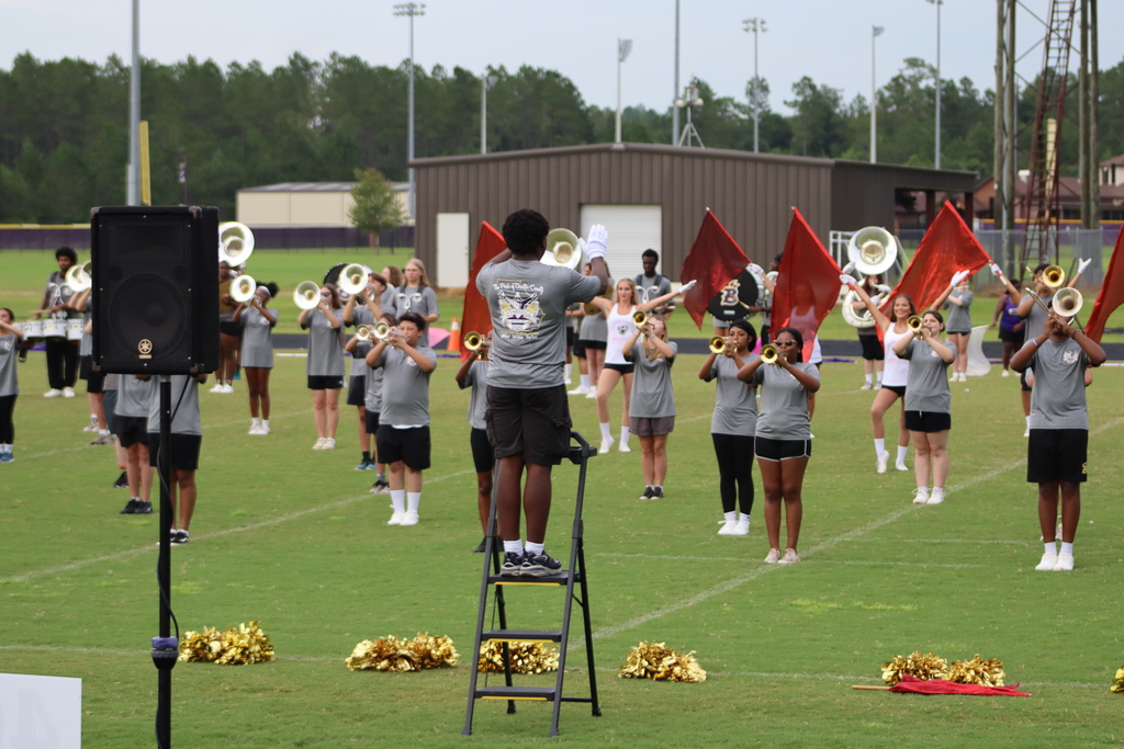 A student drum major stands on a ladder conducting the BHS Marching Band during a full ensemble rehearsal; brass instruments and red flags are raised across the field