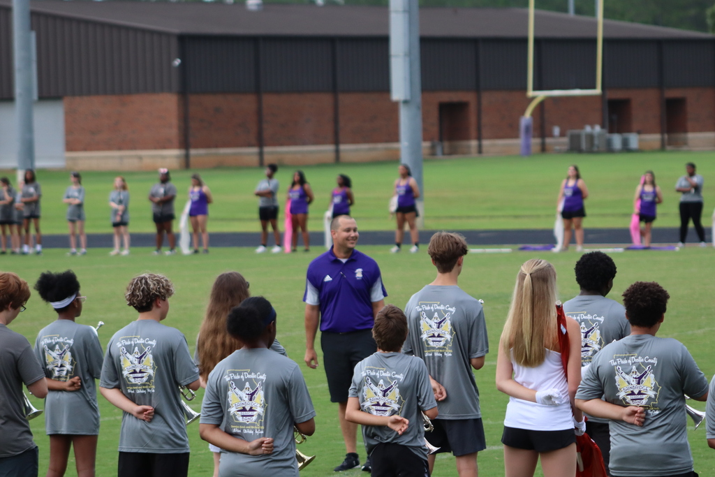 A coach smiles while addressing students on the field during BHS Marching Band Family & Friends Night, with auxiliary members lined up in the background.