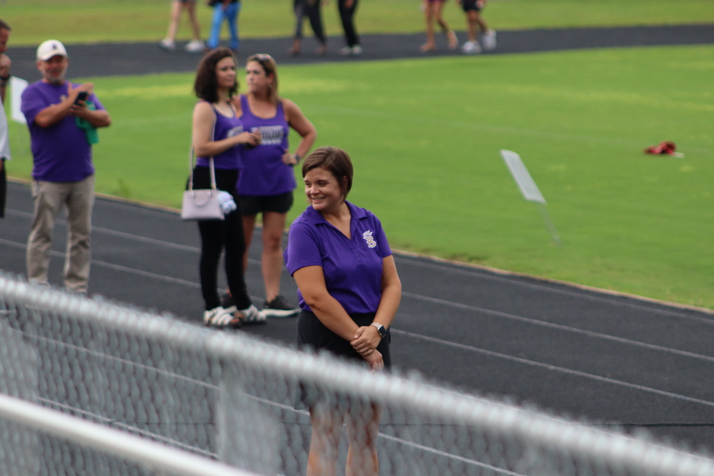 A smiling staff member stands on the track near the field, wearing a purple BHS polo, while parents and supporters look on in the background.