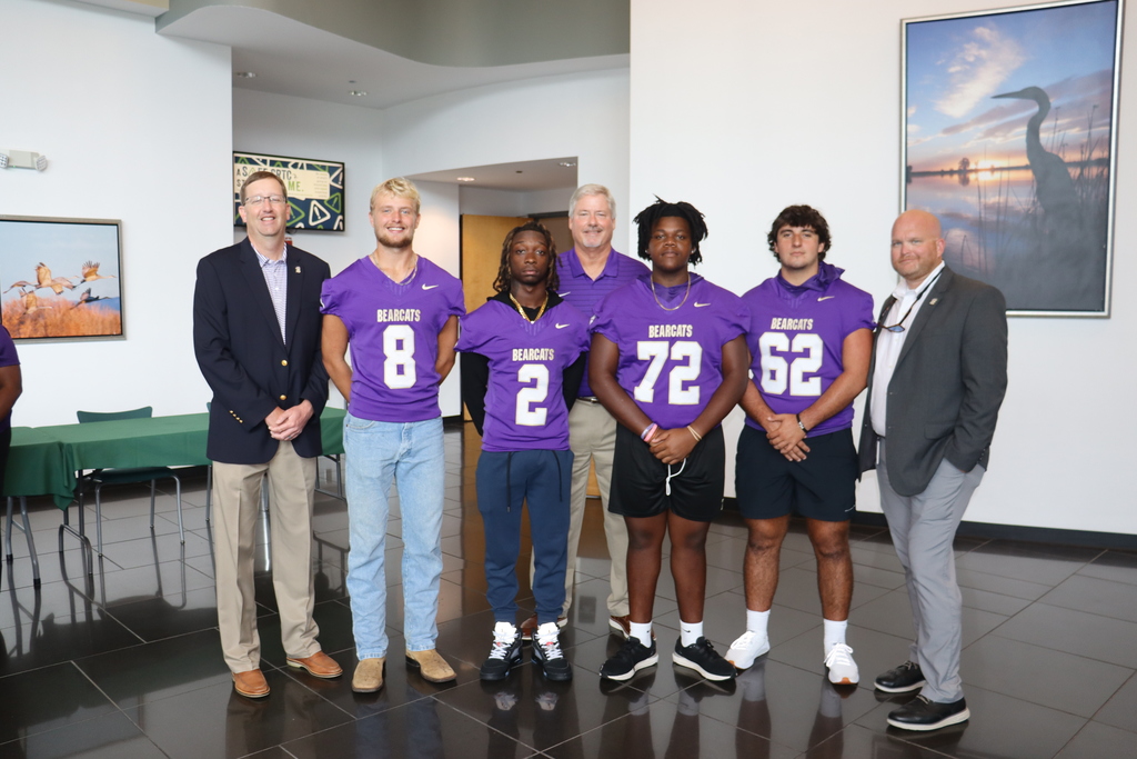 A group of Bainbridge High School football players in purple jerseys stand with school and community leaders inside a building lobby.
