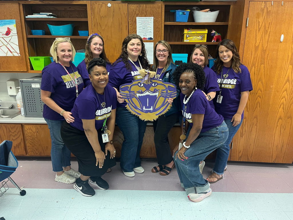 A group of eight smiling Decatur County Schools staff members wearing matching purple Bainbridge Bearcats shirts, posing together in a classroom while holding a large Bearcat mascot cutout.