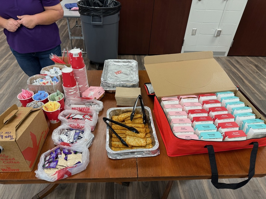 Table with various food items and supplies including small cups with packets, wrapped items, a box labeled "Thank you" with Wawa branding, fried food container with tongs, stacks of pink and blue boxes, and a red bag, with part of a person and room visible behind.