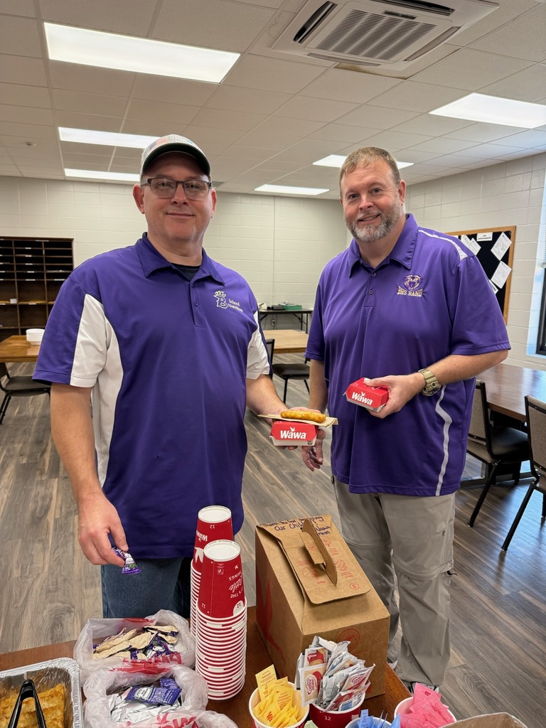 Two men indoors wearing purple shirts with logos, holding food containers, standing near tables with food and drinks, with a bulletin board behind them.