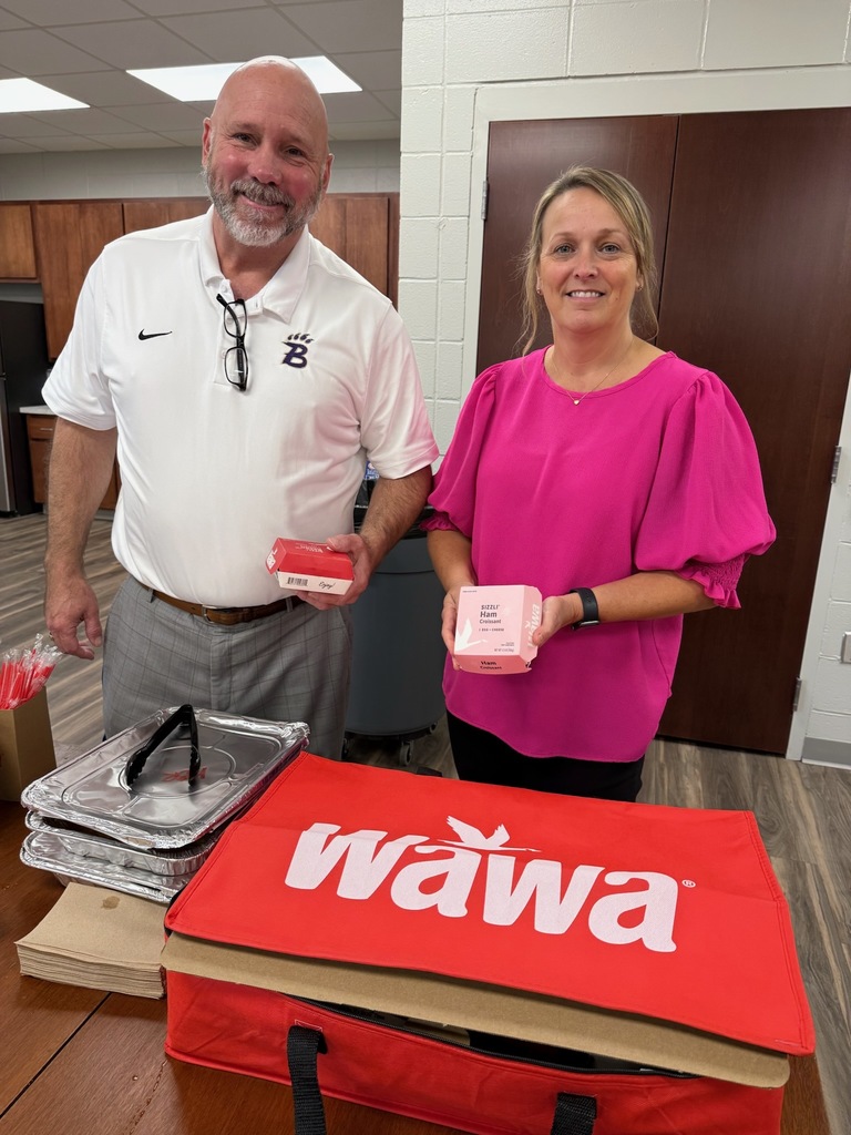 Two people indoors smiling at the camera; man in white polo with logos, woman in bright pink holding a pink box labeled "SIZZUR Ham Croissant," standing behind a table with food containers and a red Wawa bag.