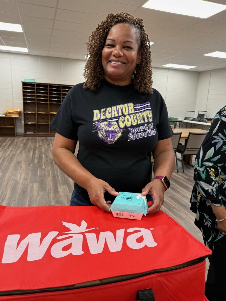 Woman indoors smiling, wearing a black "DECATUR COUNTY Board of Education" T-shirt holding a small box, with a red Wawa bag and room with tables and chairs behind her.