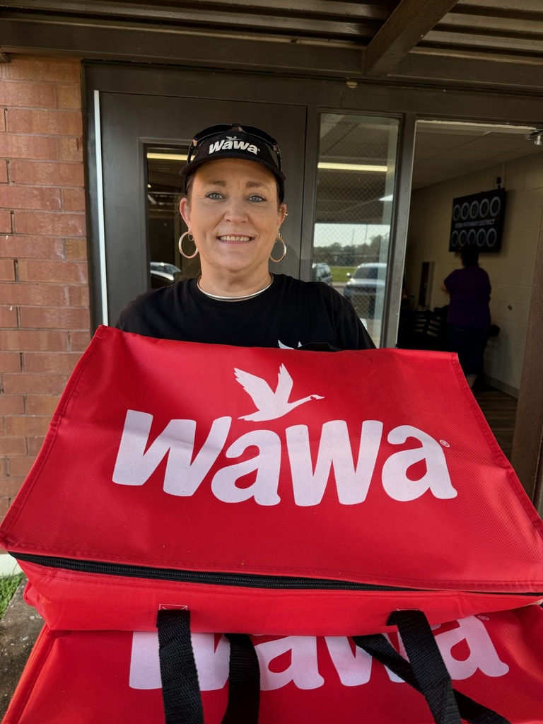 Woman standing outside holding a large red Wawa bag, wearing a black Wawa cap and shirt, with a brick wall and glass panel door behind her.