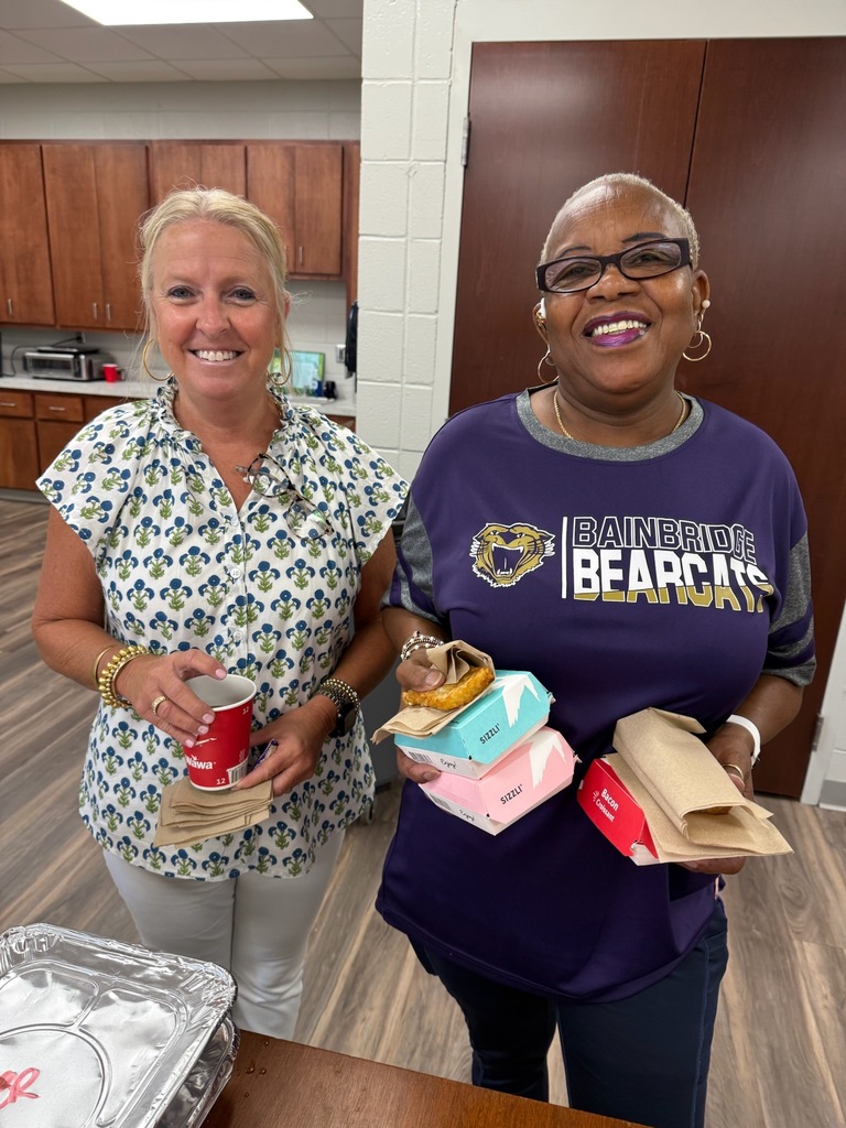 Two women indoors smiling; one in a white floral blouse holding a red cup and napkins, the other in a purple "Bainbridge Bearcats" shirt holding food containers and napkins, with kitchen cabinets in the background.
