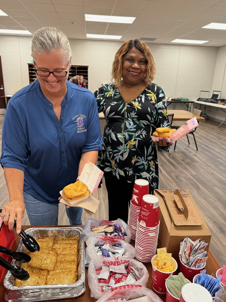 Two women standing at a table with food; one in a blue shirt with "Decatur County School Nutrition" embroidery holding a food tray, the other in a black floral top holding a tray with a pastry, in a cafeteria-like room.