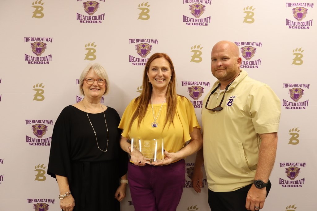 Three people stand in front of a backdrop with "THE BEARCAT WAY," "DECATUR COUNTY SCHOOLS," a purple bearcat logo, and a gold "B" logo. From left to right: an older woman with glasses and short light hair wearing black; a woman with long red hair in a yellow top and purple pants holding a glass award; and a bald man with a beard wearing a light yellow shirt with a purple "B" logo and sunglasses around his neck.