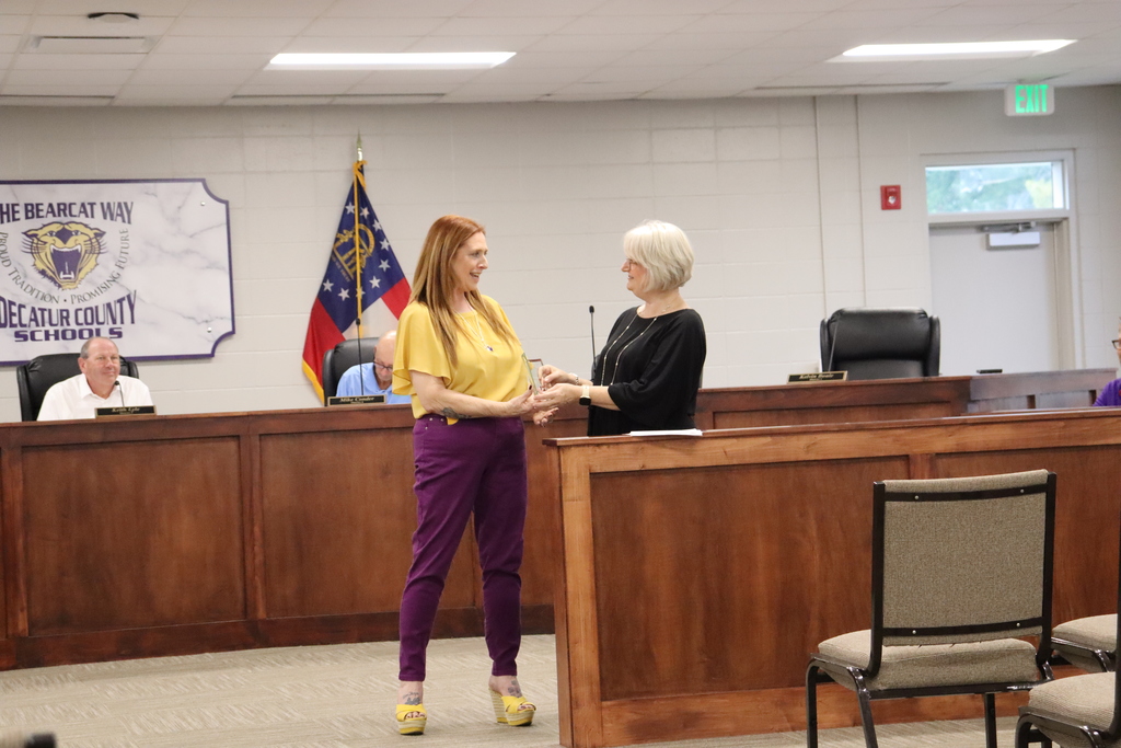 In a formal meeting room, a woman with long red hair wearing yellow and purple stands smiling, holding an award while facing a woman with short white hair in black who appears to be presenting it. Three men sit behind a wooden desk watching. The background shows a U.S. flag, a sign reading "THE BEARCAT WAY," and "DECATUR COUNTY SCHOOLS