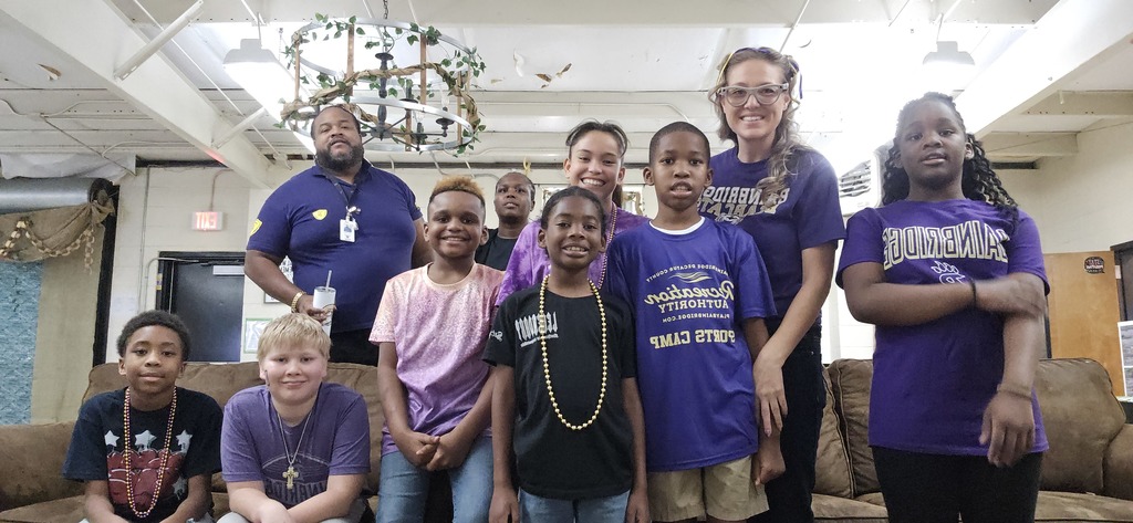 A group of children and two adults pose together indoors, smiling at the camera. Most of the group is wearing purple Bainbridge-themed shirts, with one adult in a navy polo. The setting appears to be a recreation or community center with couches, exposed ceiling beams, and a decorative hanging light fixture above.