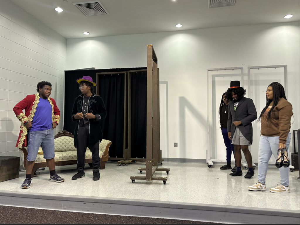 A group of students rehearses on stage in costume. One student wears a red coat with gold trim, another wears a purple hat and black jacket, while others wear jackets and top hats. The vintage couch and wooden stage frames are visible behind them.
