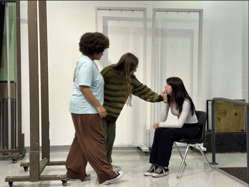 Three students perform a scene. One sits in a chair looking thoughtful while another gently touches her face. A third student stands beside them. All are dressed casually, and the wooden stage frames are positioned around them.