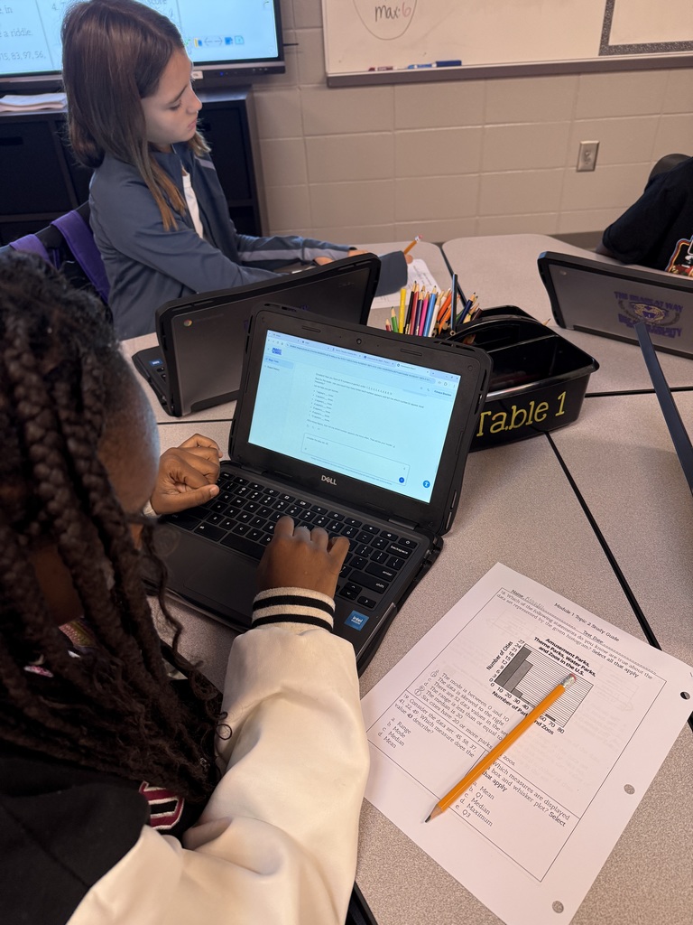 A workspace photo showing a person using a Dell laptop displaying an online quiz from "MAGIC SCHOOL," alongside a printed worksheet, a yellow pencil, and office supplies like colored pencils and markers.