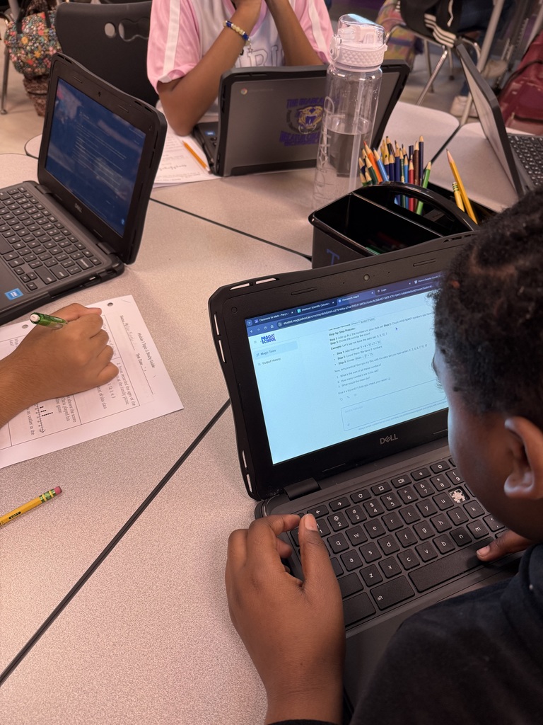 A workspace photo showing a person using a Dell laptop displaying an online quiz from "MAGIC SCHOOL," alongside a printed worksheet, a yellow pencil, and office supplies like colored pencils and markers.
