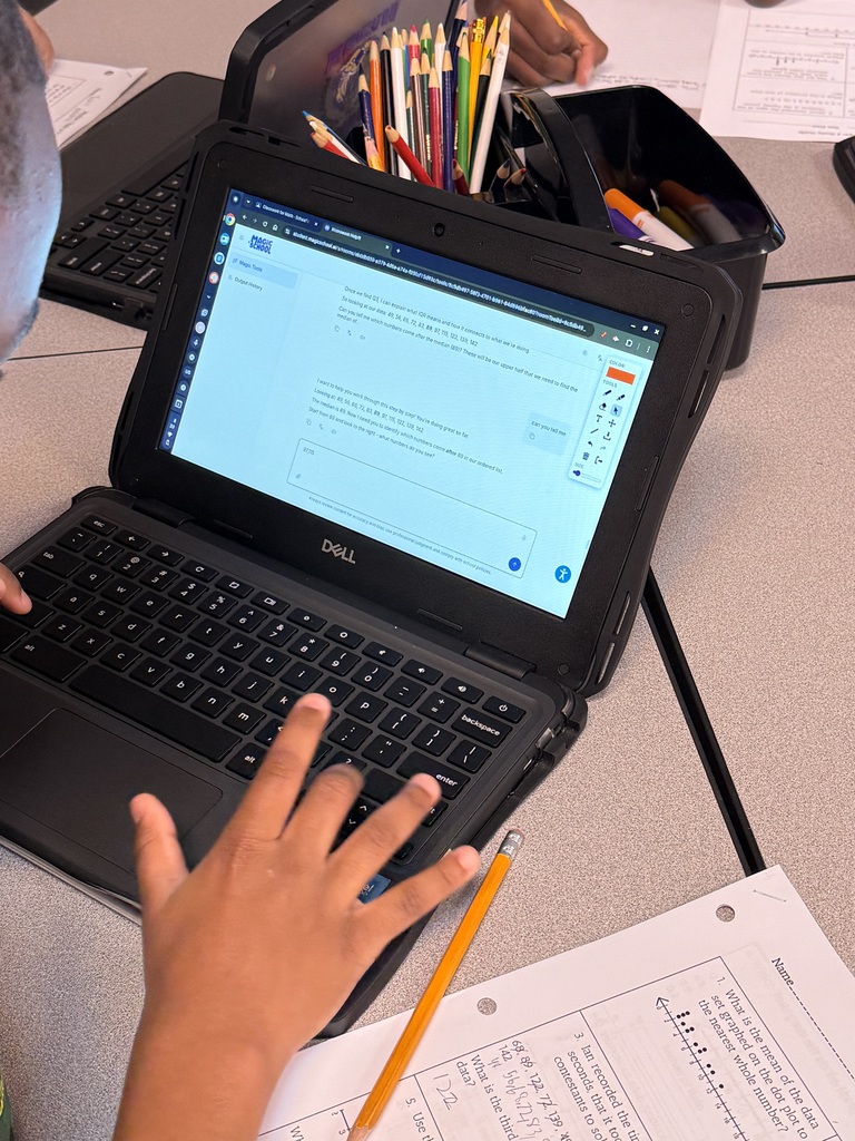 A workspace photo showing a person using a Dell laptop displaying an online quiz from "MAGIC SCHOOL," alongside a printed worksheet, a yellow pencil, and office supplies like colored pencils and markers.