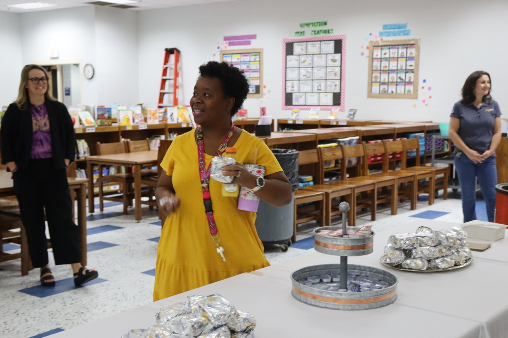 An educator in a yellow dress smiles while holding a biscuit and drink at the breakfast event inside the school library. Other staff look on in the background.