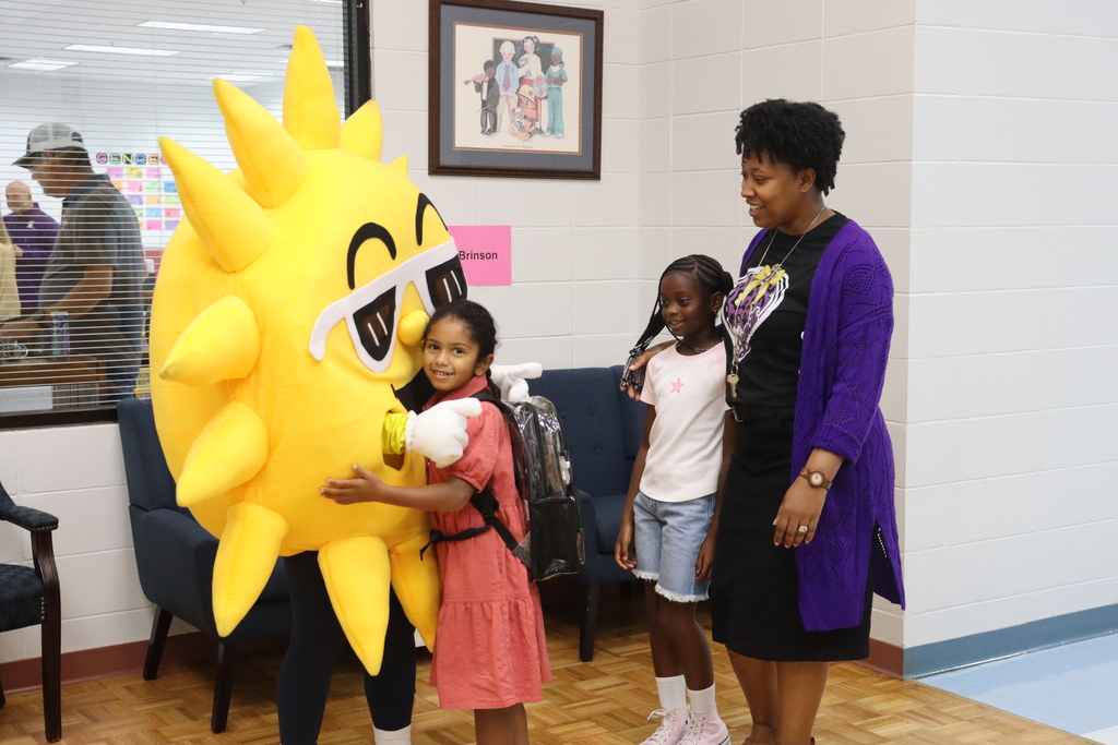 Sunny the SunStop mascot gives a big hug to a young student as another child and an adult smile nearby in the school hallway.