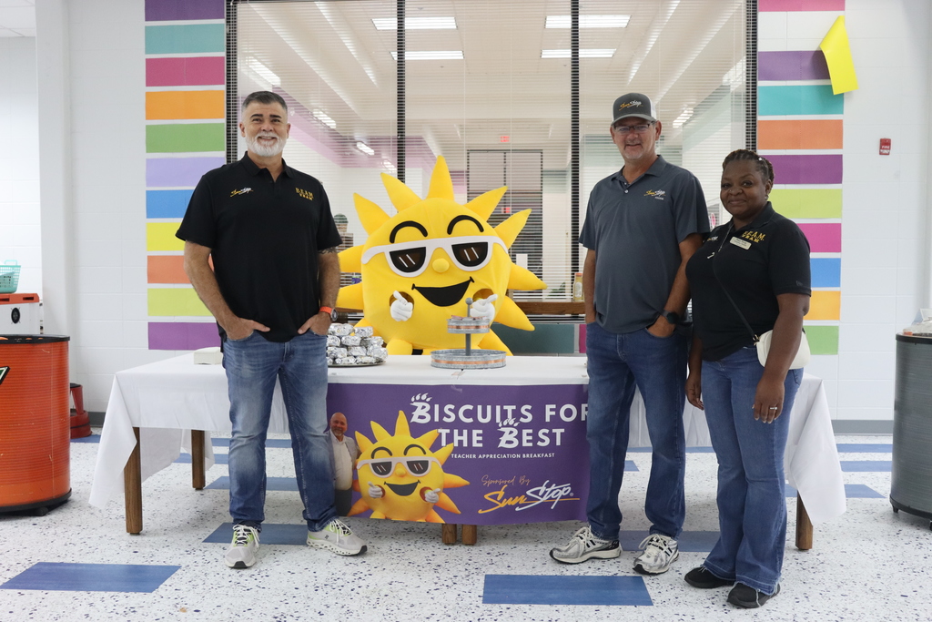 Three adults pose with Sunny the SunStop mascot behind a table decorated with a “Biscuits for the Best” banner at West Bainbridge Primary. Wrapped breakfast biscuits are stacked on the table.