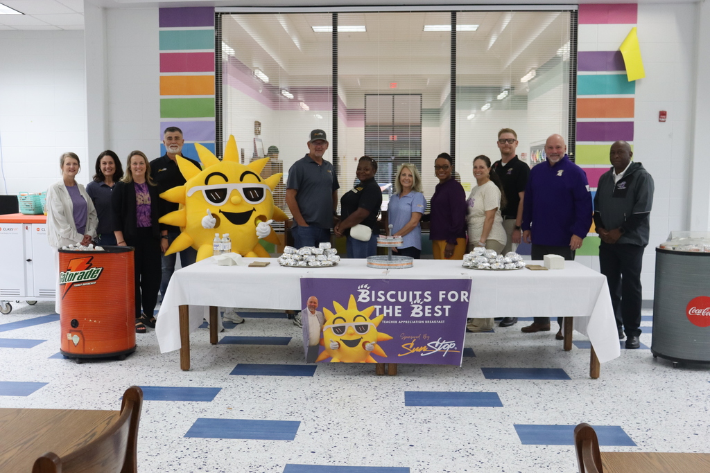 A large group including Superintendent Dr. Boyd English, district staff, and SunStop partners pose with Sunny the mascot behind a table filled with breakfast biscuits and a banner reading “Biscuits for the Best.”