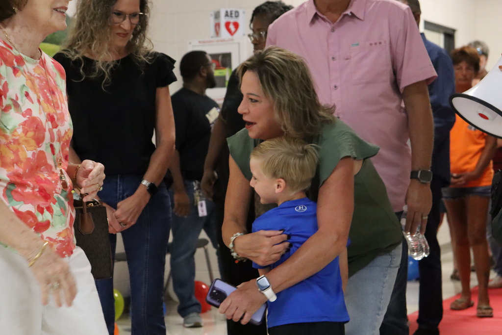 A woman crouches down to hug a young boy in a blue shirt while other adults and children walk by in a crowded school hallway.