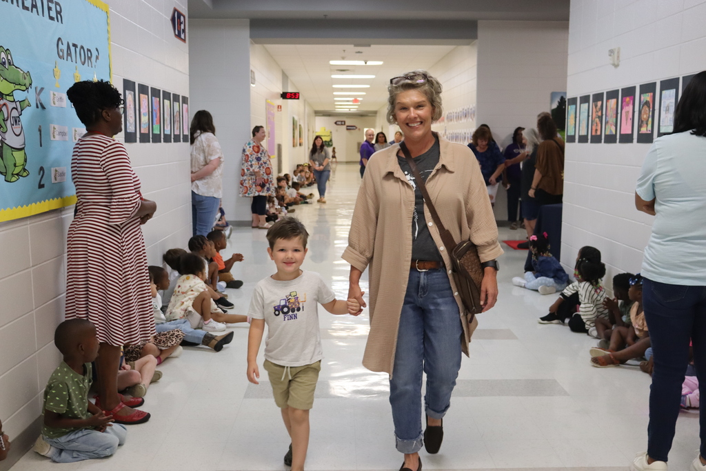 A smiling woman walks hand-in-hand with a young boy down a school hallway lined with seated students watching the grandparents' arrival.