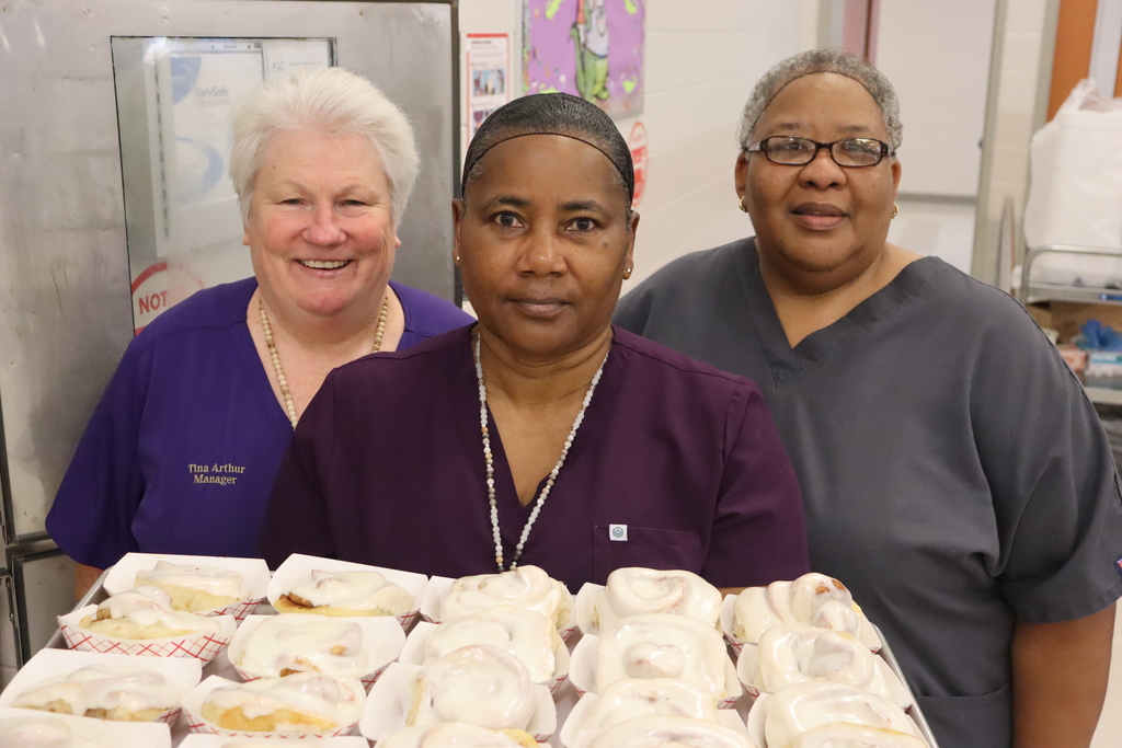 Three cafeteria staff members stand together holding trays of freshly frosted cinnamon rolls, smiling for the camera.