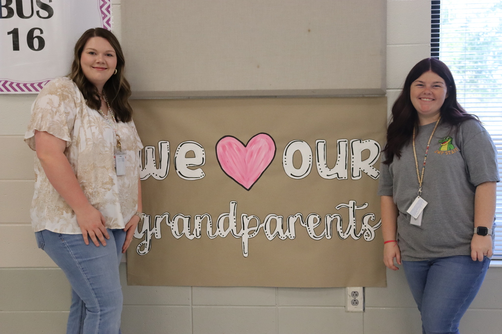 Two smiling women stand on either side of a wall display that reads "We ❤️ Our Grandparents" in large white letters with a pink heart in the center.